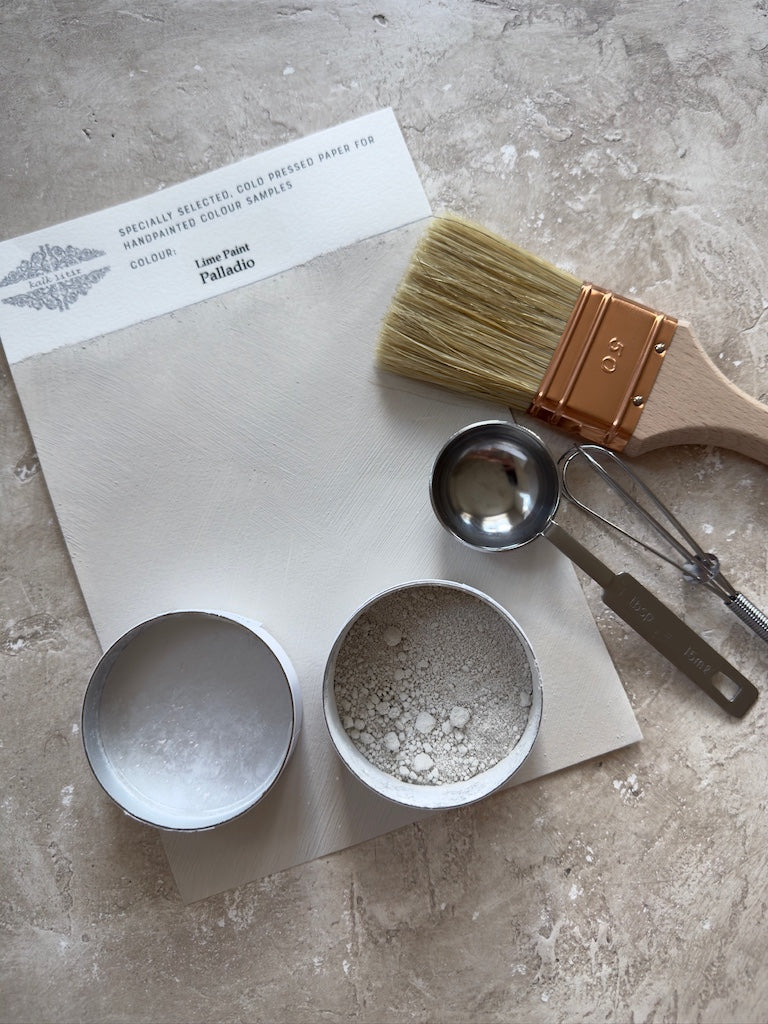 Paintbrush, measuring cups, and a small bowl on a textured surface with a paint color sample.